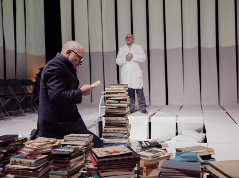 Scena teatralna. Na pierwszym planie kilka stosów książek. Wśród nich klęczy mężczyzna w płaszczu z otwartą książką w dłoniach. W tle stoi drugi mężczyzna w fartuchu lekarskim.
