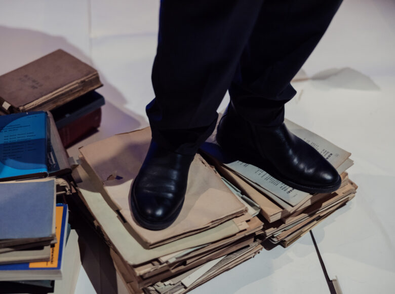 Scena teatralna. Zbliżenie na męskie nogi w czarnych eleganckich butach i eleganckich czarnych spodniach. Mężczyzna stoi na stosie teczek z dokumentami. Obok na podłodze leżą książki.