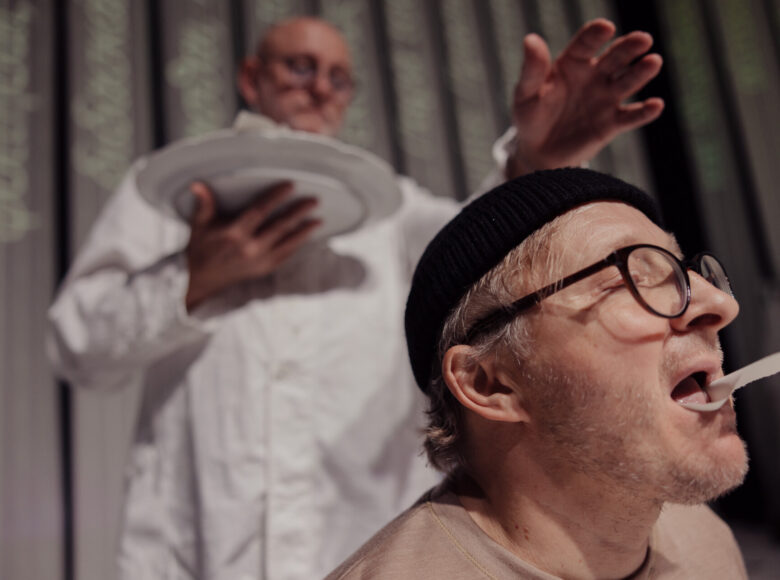 Scena teatralna. Zbliżenie na twarz mężczyzny w okularach, który trzyma na języku kawałek papieru. Za nim stoi mężczyzna w fartuchu lekarskim. W jednej ręce trzyma talerz, drugą rękę unosi w geście błogosławieństwa.