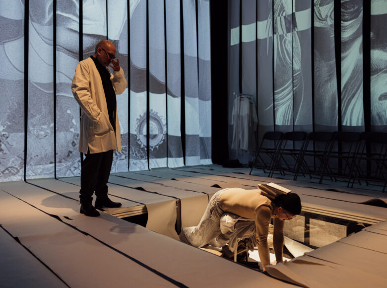 Scena teatralna. Na środku podłogi zagłębienie, w zagłębieniu postać stojąca na czworakach. Na jej karku leży kilka książek. Obok na podłodze stoi mężczyzna w fartuchu lekarskim.