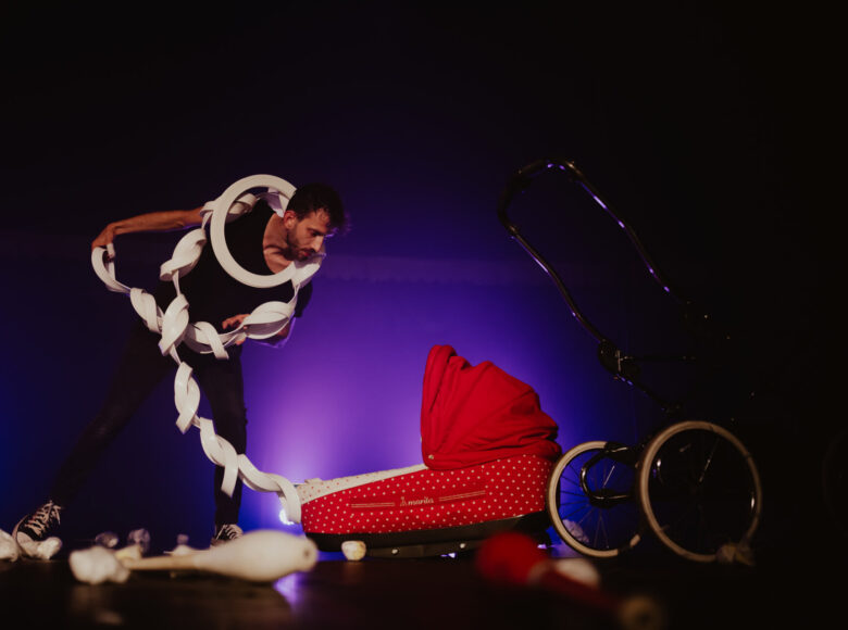 Zdjęcie przedstawia mężczyznę, który znajduje się na scenie. Na podłodze leży rozłożony na części czerwony wózek dziecięcy. Mężczyzna pochyla się nad gondolą. Na szyi ma białą obręcz, w rękach trzyma biały sznur powiązanych ze sobą pieluch.