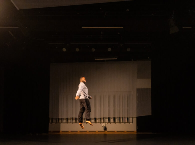 Kolorowa fotografia. Na zdjęciu mężczyzna uchwycony w trakcie wysokiego podskoku. Ubrany jest w jasną koszulę z długimi rękawami i długie ciemne spodnie. Ma bose stopy.