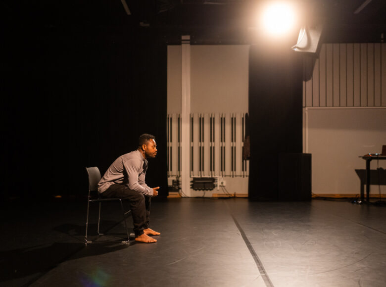 Kolorowa fotografia. Na zdjęciu mężczyzna siedzi na krześle. Ubrany jest w koszule i długie spodnie. Ma bose stopy. Patrzy przed siebie.