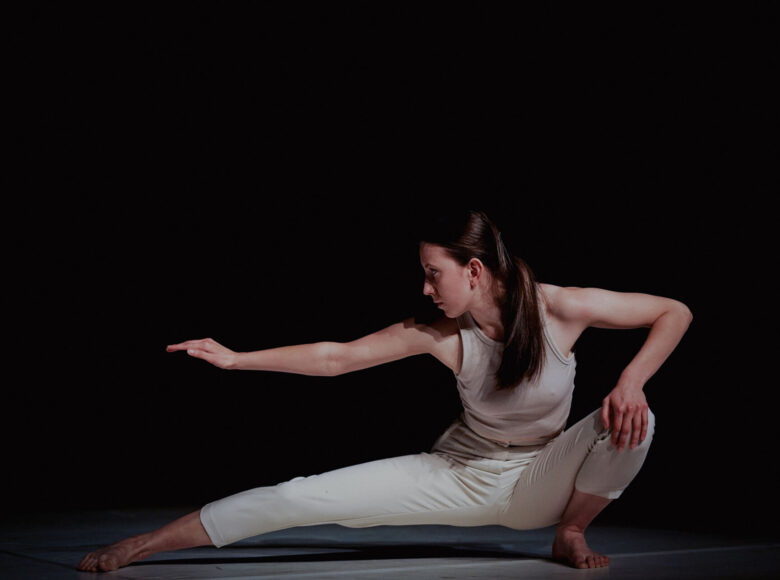 Kolorowa fotografia. Kobieta ubrana w białe spodnie i top. Kuca z prawą nogą i ręką wyprostowaną w bok. Lewą dłoń opiera na lewym kolanie. Spogląda w bok.