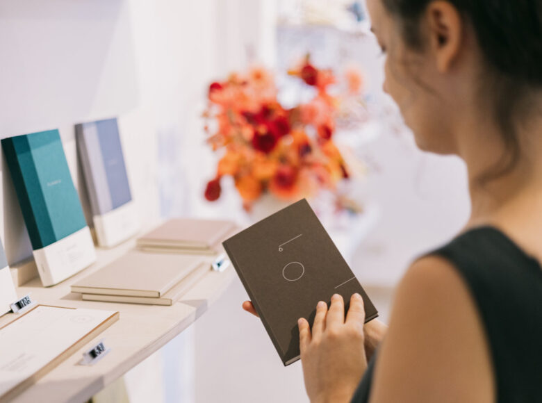 Zdjęcie przedstawia kobietę, która stoi przed sklepową półka, w ręce trzyma notes z ciemna okładką.