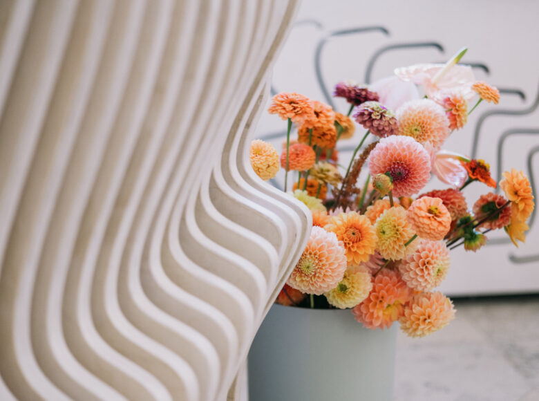 Zdjęcie przedstawia drewnianą, pofalowaną konstrukcję. obok niej stoi biały wazon z pomarańczowymi kwiatami.