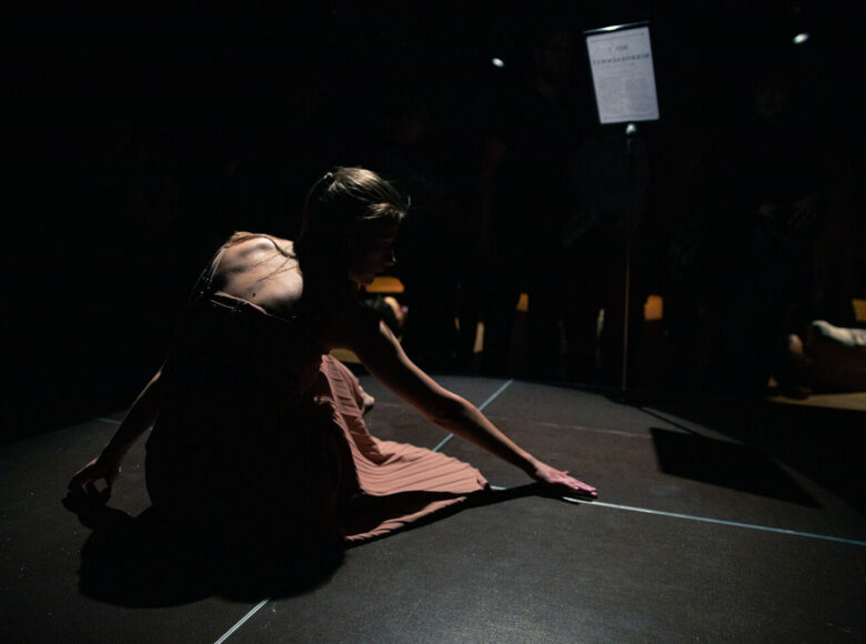 Kolorowa fotografia. Na scenie siedzi kobieta. Jej sylwetkę lekko oświetla światło. Ubrana jest w zwiewną sukienkę. Ręką gładzi podłogę.