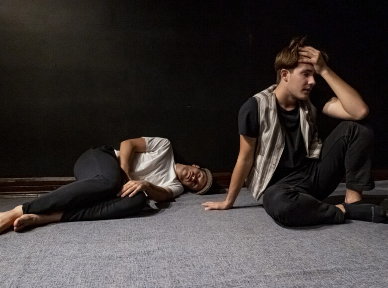 Przedstawienie teatralne. Na podłodze dwóch młodych mężczyzn. jeden siedzi, drugi leży. Za nimi czarne tło.