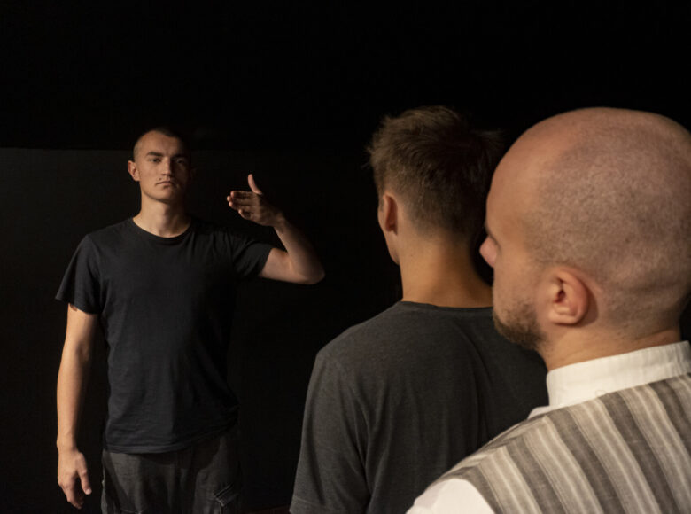 Przedstawienie teatralne. Na scenie stoi trzech mężczyzn, jeden przodem do widza a dwaj tylem. Za nimi czarne tło.