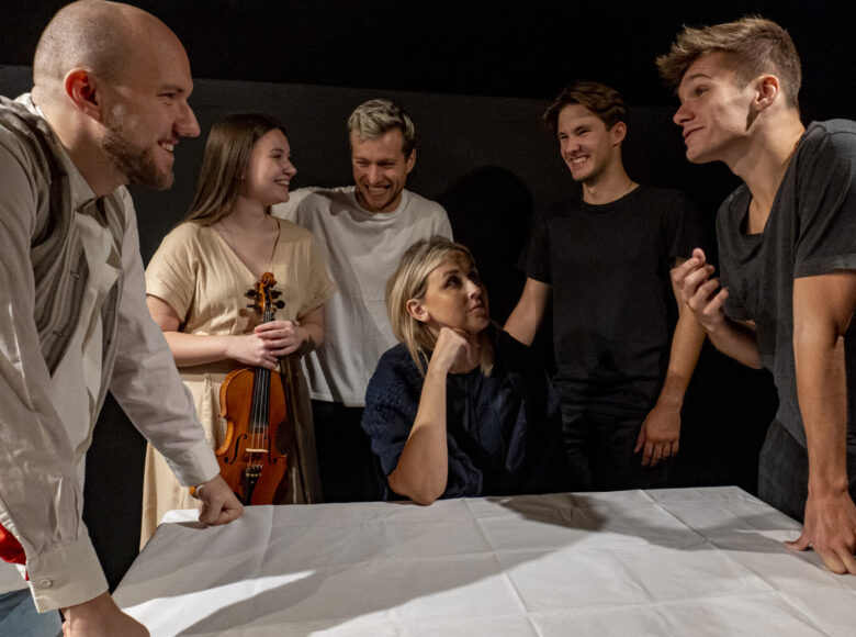 Kobieta siedzi przy stole. Wokół niej stoi grupa roześmianych osób - dziewczyna i czterech chłopaków. Dziewczyna trzyma w ręku skrzypce.