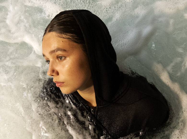 Kolorowa fotografia. Kobieta ubrana w czarną bluzę z kapturem. Kobieta jest w wodzie, otacza ją piana.