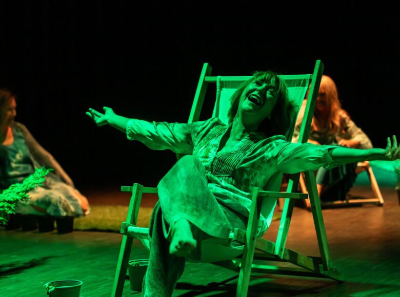 Kobieta siedzi na plażowym leżaku, rozkłada szeroko ręce, jej twarz wyraża radość. Na scenę pada zielone światło.