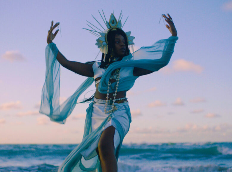 Kobieta stojąca na tle morza w porze zachodu. Kobieta w biodrach opasana białym pareo, na ramionach ma lekkie szale i naszyjnik z muszli, na głowie zdobną formę korony. Ręce ma uniesione nad głowę w geście przypominającym taniec.