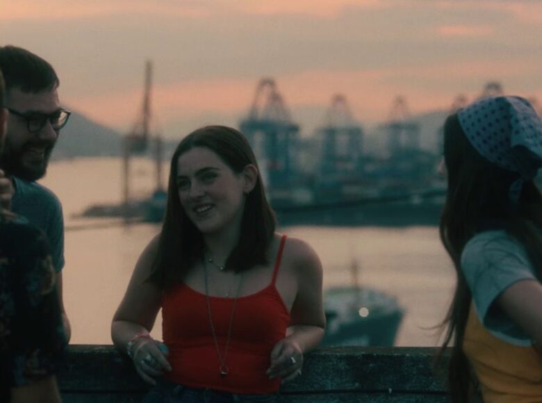 Kadr z filmu. Na tle rozmytego portowego krajobrazu stoją trzy dziewczyny i dwóch chłopaków. Uśmiechają się, rozmawiają ze sobą.