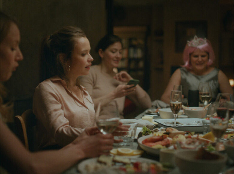 Kadr z filmu. Za stołem wypełnionym jedzeniem i kieliszkami siedzą cztery młode kobiety. Jedna z nich ma na głowie różową perukę i diadem.