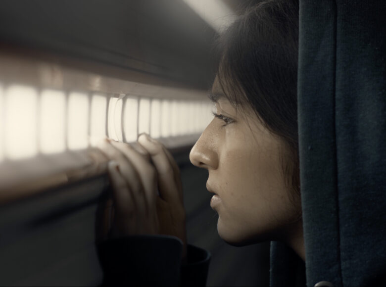 Kadr z filmu. Zbliżenie na twarz młodej dziewczyny w kapturze. Stoi przed podłużną kratką, przez którą wpada światło. Dziewczyna trzyma dłonie na jej dolnej krawędzi i patrzy przez nią.