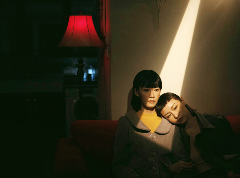 Na zdjęciu dwie młode kobiety o azjatyckich rysach twarzy. Siedzą oparte o ścianę, jedna opiera twrz na ramieniu drugiej. Twarze mają oświetlone ostrym snopem słońca, pozostała część zdjęcia w cieniu.