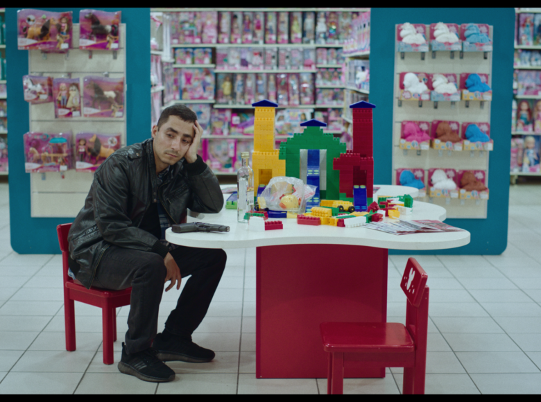 Kadr z filmu. Wnętrze dużego sklepu z zabawkami. Na środku przy stoliku dla dzieci siedzi mężczyzna z ponurą miną. Głowę podpiera ręką. Obok niego na stoliku pistolet.