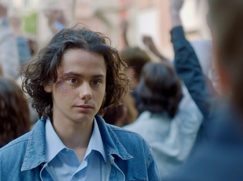 Kadr z filmu. Zbliżenie na młodego chłopaka stojącego w tłumie na zewnątrz. Chłopak jest w dżinsowej kurtce, ma długie włosy i siniak pod okiem.