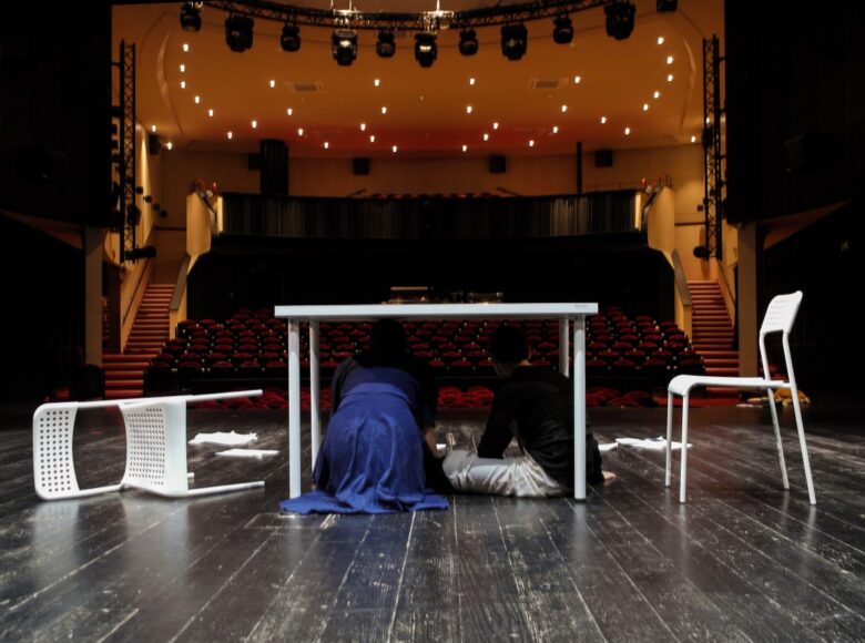 Na zdjęciu dwie osoby kucają pod białym stołem, odwrócone tyłem do kadru. Stół znajduje się na pustej scenie teatralnej, obok niego dwa przewrócone białe krzesła.