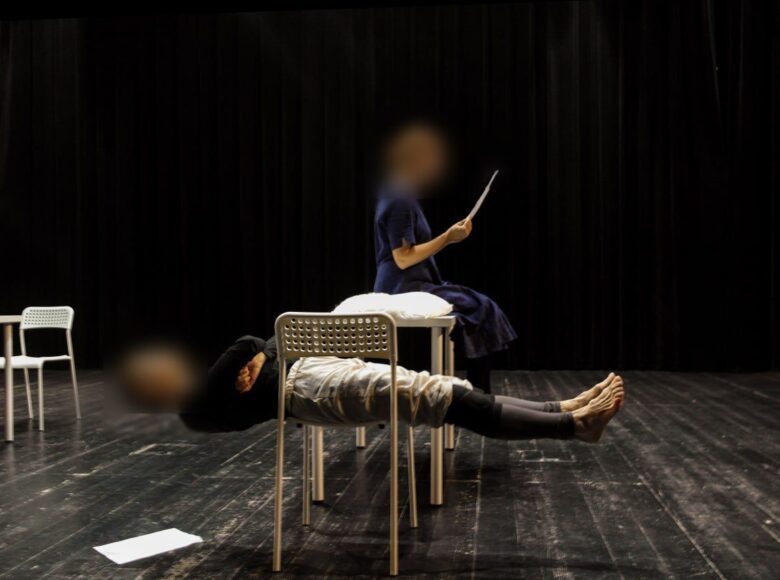 Na kolorowym zdjęciu jedna osoba leży na krześle, ma wyprostowane nogi i ręce założone z przodu na krzyż. Druga osoba siedzi przy białym stole na krześle i czyta. Całość na pustej scenie teatralnej.