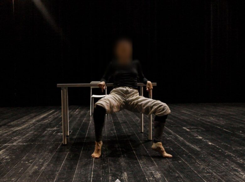 Na zdjęciu osoba opierająca się łokciami o stół. Ma bose stopy, stoi na palcach, w figurze przypominającej ćwiczenie taneczne.