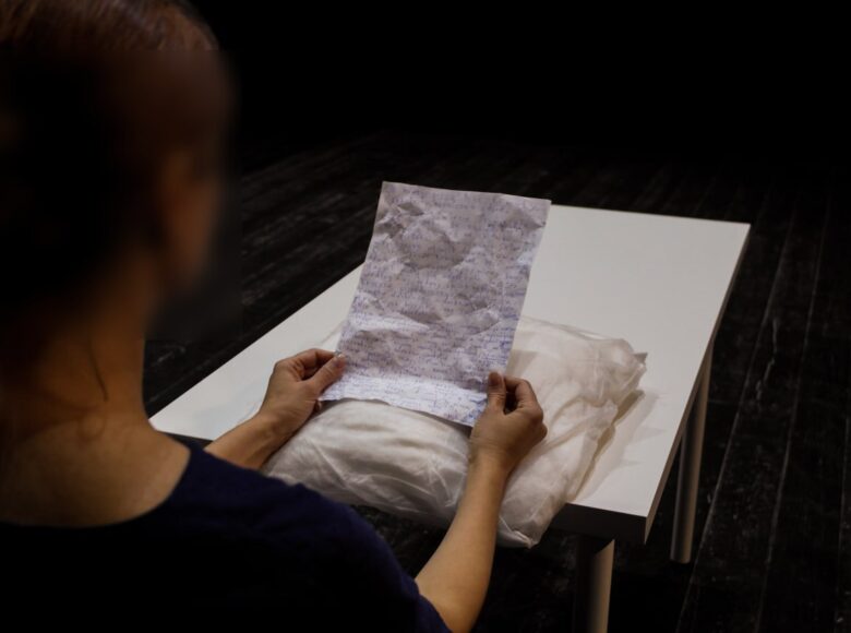 Na zdjęciu osoba siedząca tyłem do kadru. W dłoniach trzyma pomiętą, zapisaną kartkę, na kolanach ma poduszkę opartą o biały stół stojący przed sobą.