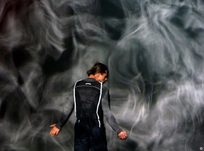 Kolorowa fotografia. Mężczyzna stoi twarzą do ściany. Ręce trzyma lekko uniesione na wysokości bioder. Na ścianę wyświetlana jest projekcja, która sprawia, że ściana wygląda jak falująca tafla wody.