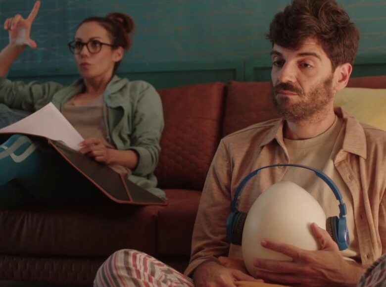 Kadr z filmu. Wnętrze. Na podłodze przed kanapą siedzi po turecku mężczyzna. Do brzucha przyciska duże jajo, które ma założone niebieskie słuchawki. Na kanapie za nim siedzi kobieta, która wyciąga nogi na pufie, na kolanach trzyma teczkę z papierami.