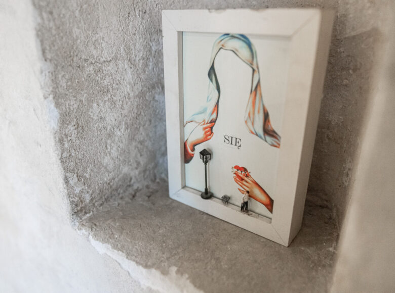 Kolorowe zdjęcie przedstawia prostokątną białą ramkę za zdjęcie z narysowanym wizerunkiem Matki Boskiej, która trzyma w dłoni serce. Na dole ramki stoi miniaturowa figurka latarni ulicznej, psa i postaci ludzkiej. Na środku obrazka napis "Się".