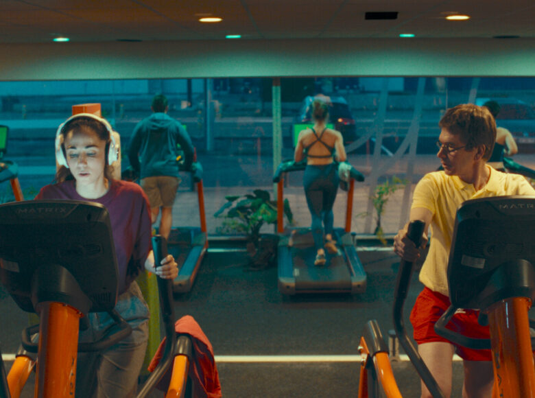 Kadr z filmu. Wnętrze siłowni. Na bieżniach i orbitrekach ćwiczą ludzie, na pierwszym planie dziewczyna i mężczyzna. Ona ma twarz oświetloną wyświetlaczem urządzenia, na którym ćwiczy, on patrzy na nią.