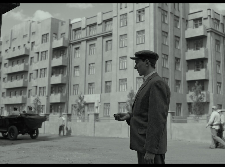 Kadr z filmu. Dwudziestolecie międzywojenne w dwudziestym wieku. Wiosenny dzień. Miejska ulica z domami. Na pierwszym planie mężczyzna w garniturze i kaszkiecie. W tle spacerujący ludzie i staroświecki samochód.