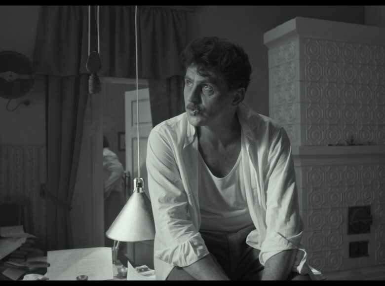 Kadr z filmu. Dwudziestolecie międzywojenne dwudziestego wieku. Wnętrze mieszkania. Na pierwszym planie na biurku siedzi mężczyzna w rozpiętej koszuli, podkoszulku i spodniach. Za nim wnętrze gabinetu, pod ścianą krzesło, na krześle leżą książki. W rogu duży kaflowy piec. W tle drzwi do drugiego pokoju, za drzwiami kobieta odwrócona tyłem.