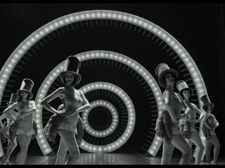 Czarno-biały kadr z filmu. Na scenie znajdują się skąpo ubrane tancerki. Na głowach nosza cylindry. Za nimi znajduje się spiralna dekoracja ze światełkami.