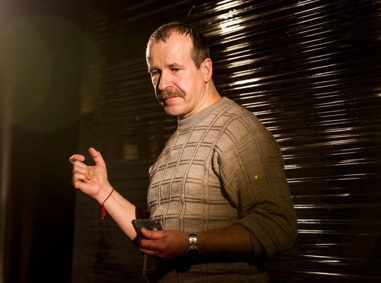 Kolorowe zdjęcie mężczyzny w średnim wieku, który stoi na tle fakturowanej w paski ciemnej ściany. Mężczyzna ma na sobie jasny sweter i wąsy, patrzy na kasetę magnetofonową trzymaną w dłoni.