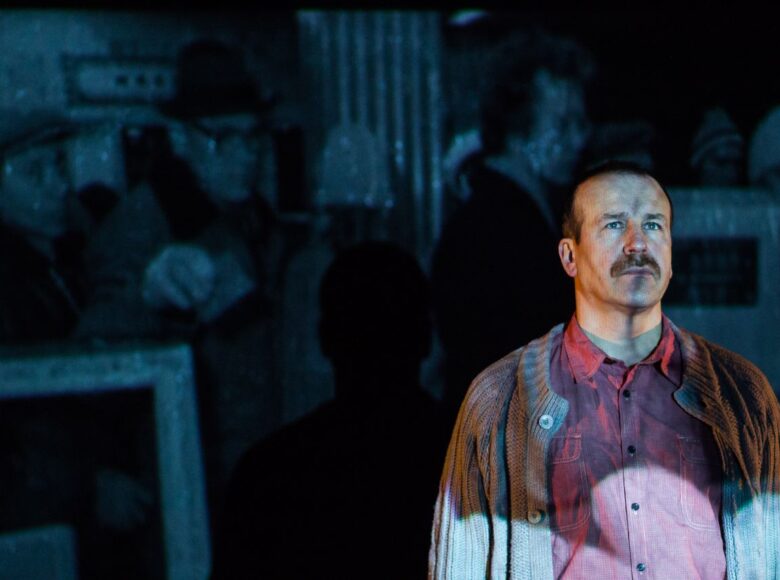 Kolorowe zdjęcie mężczyzny w średnim wieku. Ubrany jest w koszulę i gruby rozpięty sweter. Stoi na scenie oświetlony nieregularnym światłem i patrzy przed siebie. W tle na ścianie widać niewyraźne obrazy z rzutnika.