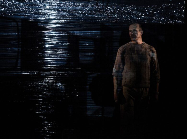 Kolorowe zdjęcie przedstawia mężczyznę w średnim wieku ubranego w gruby sweter. Stoi on na ciemnej scenie za ścianą przezroczystej folii.