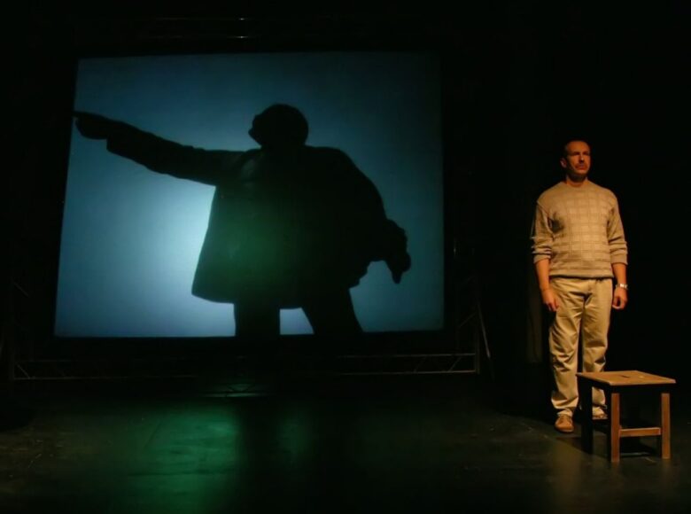 Kolorowe zdjęcie przedstawia mężczyznę stojącego po prawej stronie sceny, przed nim stoi drewniany stołek. W tle na ścianie widać cień sylwetki mężczyzny stojącego bokiem z uniesioną do góry ręką.