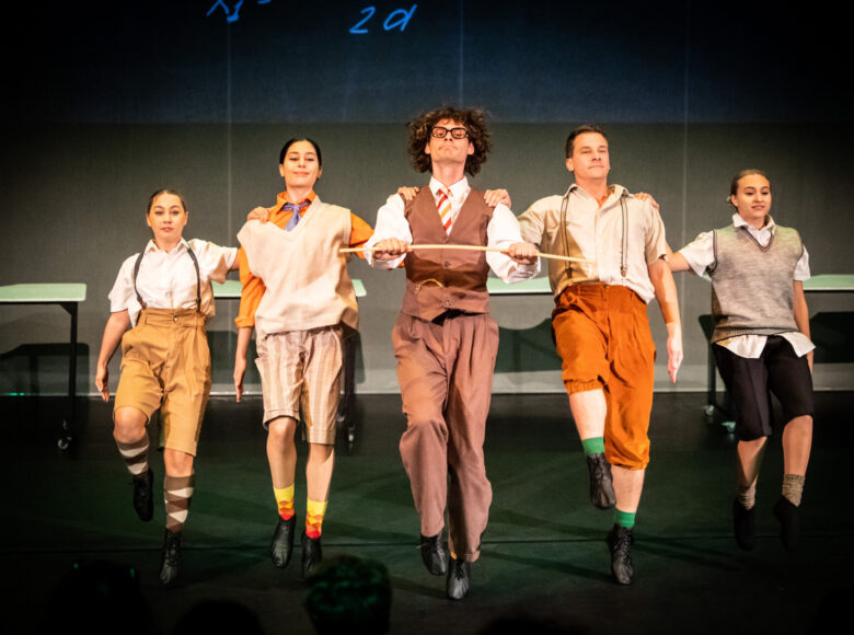 Kolorowa fotografia. Pięć osób stoi w szeregu, trzymając jedną rękę na ramieniu osoby obok. Wszyscy ubrani są w różne, kolorowe mundurki. Wyglądają jak uczniowie.