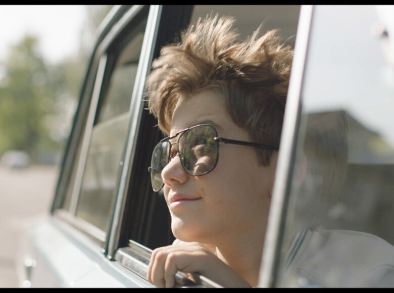 Na zdjęciu twarz chłopca wyglądającego z samochodu. Chłopiec ma okulary słoneczne. W tle droga i drzewa.