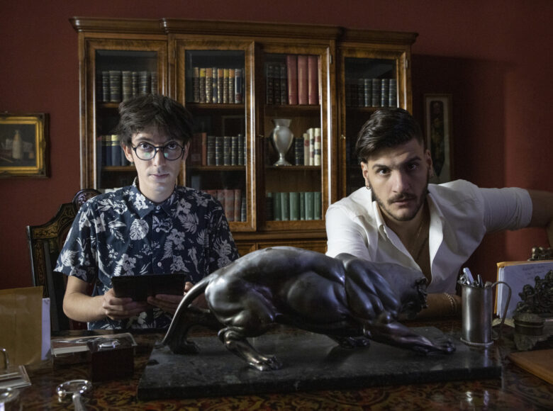 Kadr z filmu. Wnętrze eleganckiego gabinetu urządzonego w starym stylu. Na pierwszym planie za biurkiem siedzi młody mężczyzna i chłopak, ubrani w koszule. Za nimi półki z książkami.