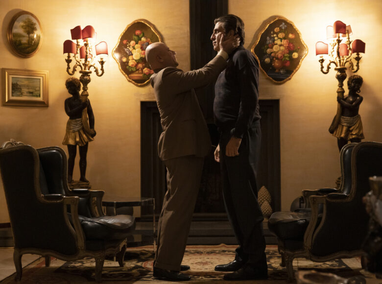 Fotos z filmu. Pokój oświetlony ciepłym światłem. Na środku dwóch mężczyzn, stojących naprzeciwko siebie. Jeden obejmuje dłońmi twarz drugiego. Za nimi kominek, po obu stronach kominka dwie rzeźby i dwa staroświeckie fotele. Na ścianach obrazy, na podłodze dywan.