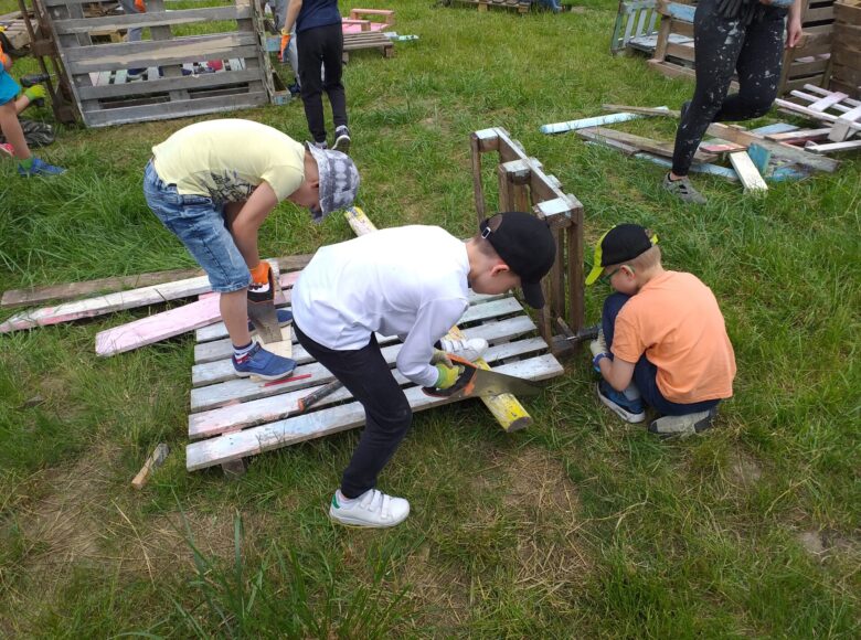 Na fotografii zajęcia majsterkowania dla dzieci na otwartym powietrzu, na trawie. Kilku chłopców tnie piłami drewniane deski.