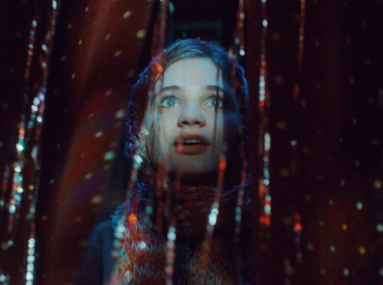 Na ciemnym tle zbliżenie na zachwyconą twarz dziewczynki. Dookoła dziewczynki zwieszaną się z sufitu błyszczące taśmy.