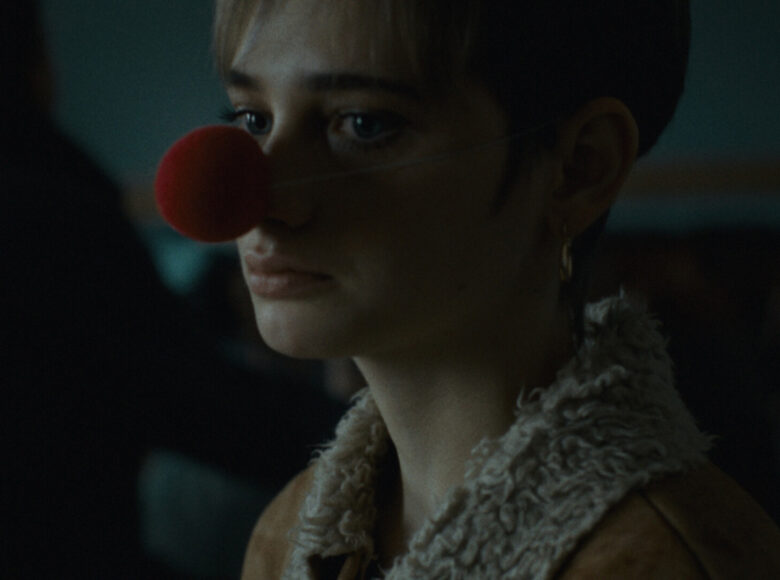 Zbliżenie na twarz kobiety. Kobieta ma przyczepiony do twarzy czerwony nos klauna. Za nią ciemne tło.