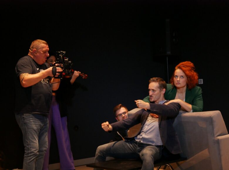 Zdjęcie przedstawia mężczyznę, który siedzi na kanapie, wyciąga przed siebie obie ręce i udaje, że kieruje samochodem. Za nim siedzi kobieta i kładzie mu ręce na ramionach. Na scenie znajduje się również mężczyzna z kamerą.