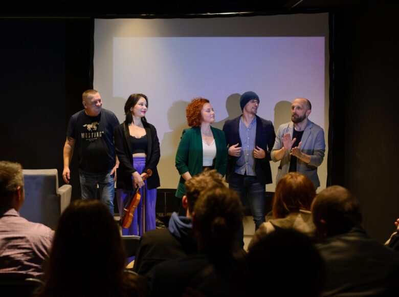 Zdjęcie przedstawia grupę aktorów, którzy soją na scenie. Za nimi znajduje się biały ekran.