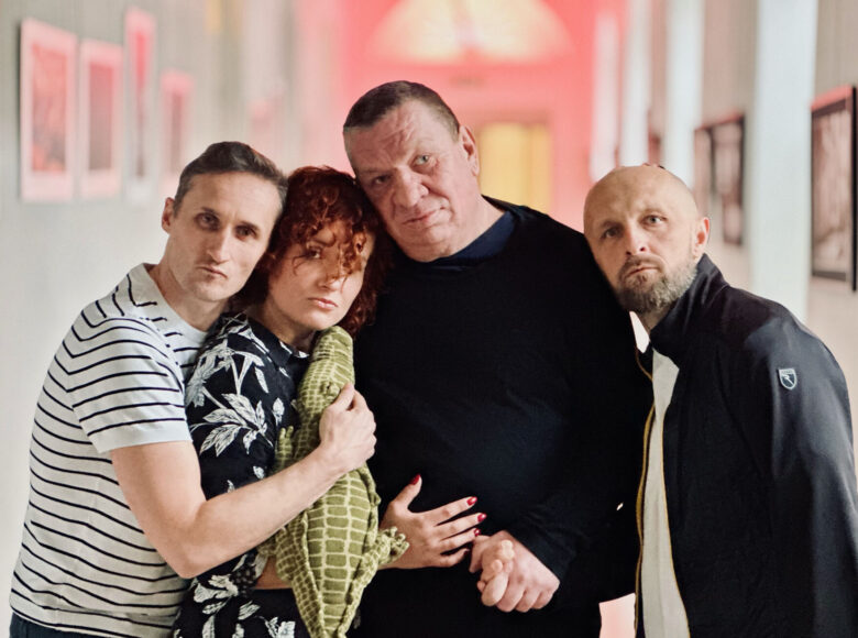 Fotografia. W pomieszczeniu kobieta i trzech mężczyzn, z bardzo poważnymi minami. Obejmują się. Jeden z mężczyzn trzyma w ręku pluszowego krokodyla. Za nimi widać obrazy na ścianach.