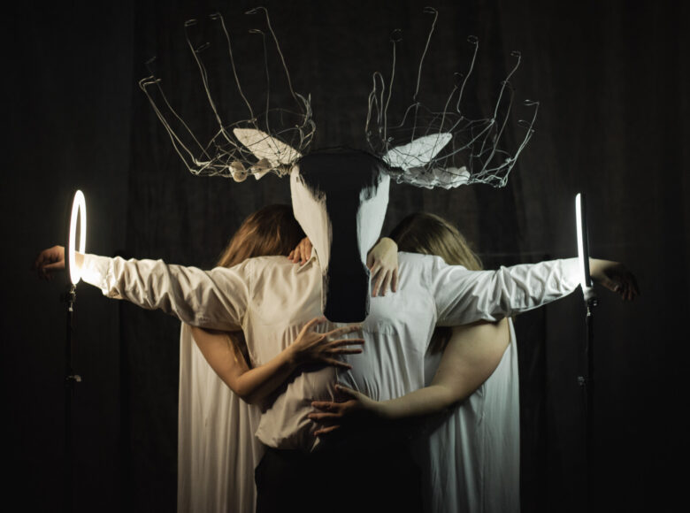 Na zdjęciu postać, na twarzy ma dużą, papierową maskę przypominającą jelenia, w której poroże jest wykonane z drutu. Ręce ma rozłożóne szeroko, a dłonie znajdują się w dwóch świecących obręczach. Po obu stronach stoją osoby o blond włosach, które obejmują główną postać, ale twarze mają schowane za nią.