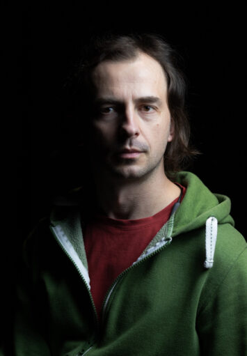 Zdjecie przedstawia portret mężczyzny w zielonej bluzie. Światło pada na połowę twarzy, druga połowa jest zaciemniona.